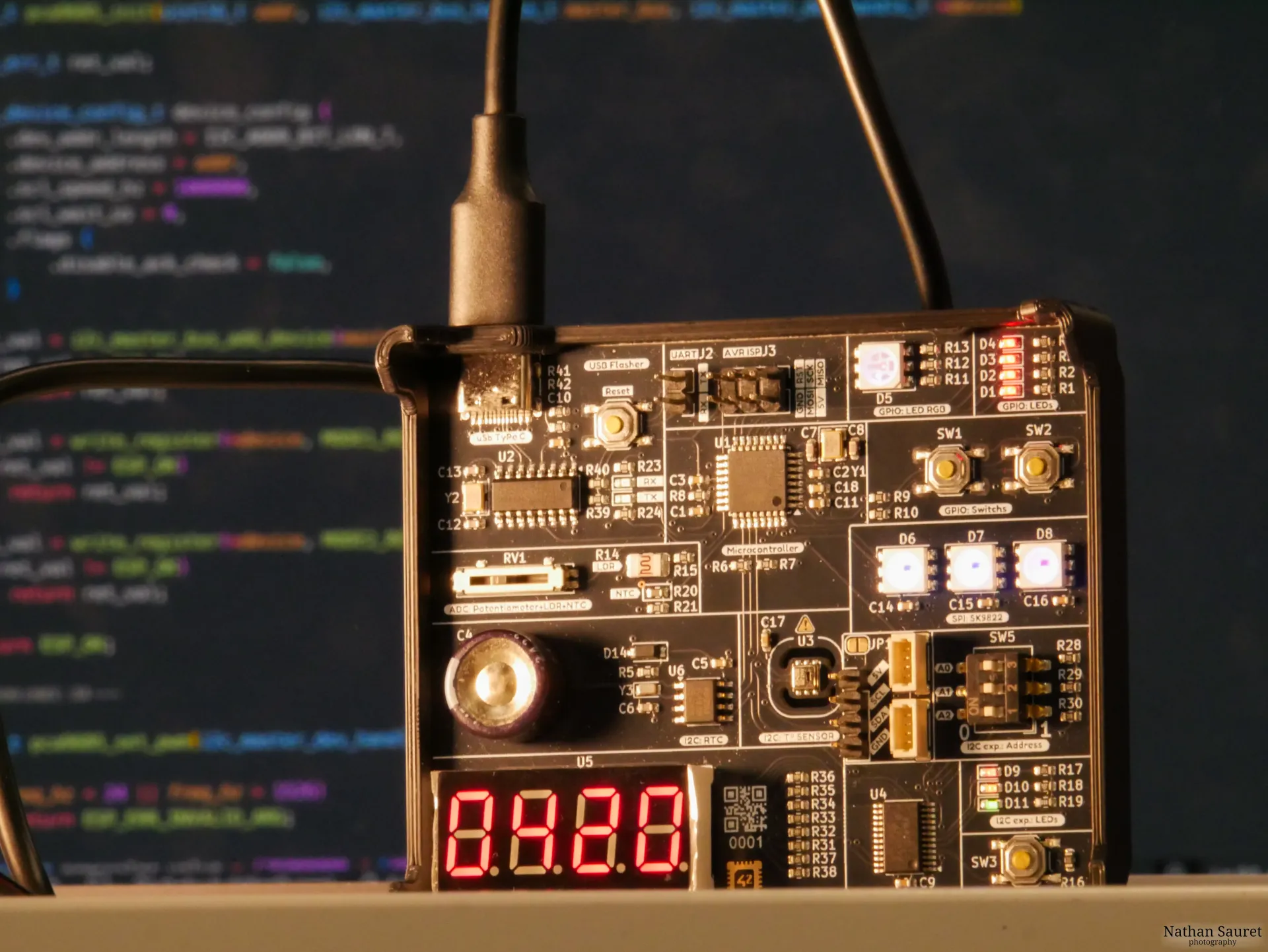 Close-up of a 42chips embedded-C piscine circuit board, its display reading “0420”, with a code editor blurred in the background.
