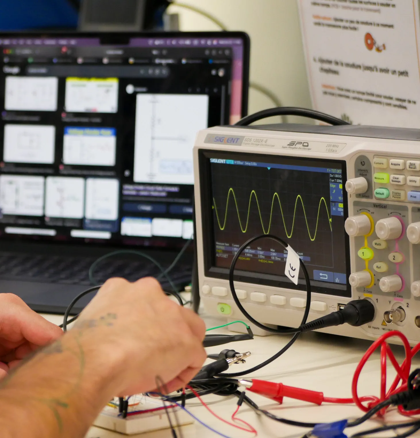Hands probing a breadboard circuit, an oscilloscope showing a clean sine wave behind them.